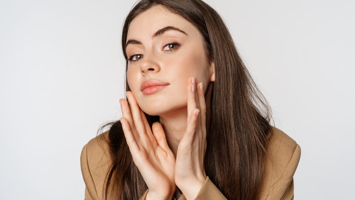 Beauty and skin care. Close up portrait of beautiful woman looking in mirror and touching her clear glowing face, standing over white background.