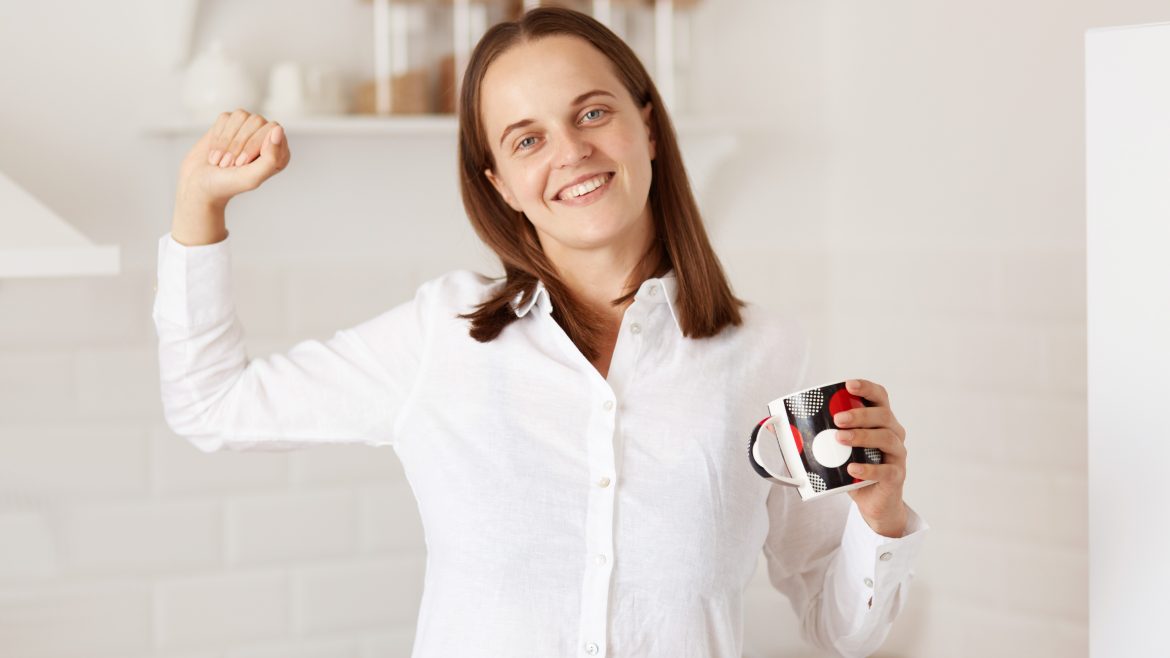 Portrait of happy woman standing with raised arm, holding cup with tea, stretching hands after waking up, looking at camera with with charming smile, wearing white casual style shirt.